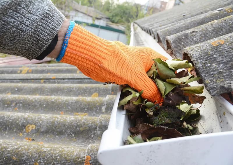 Orange-gloved hand removing leaves from a gutter beside roof tiles