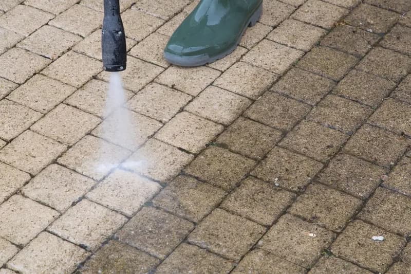Pressure washer jet lifting dirt from block paving beside a worker's boots